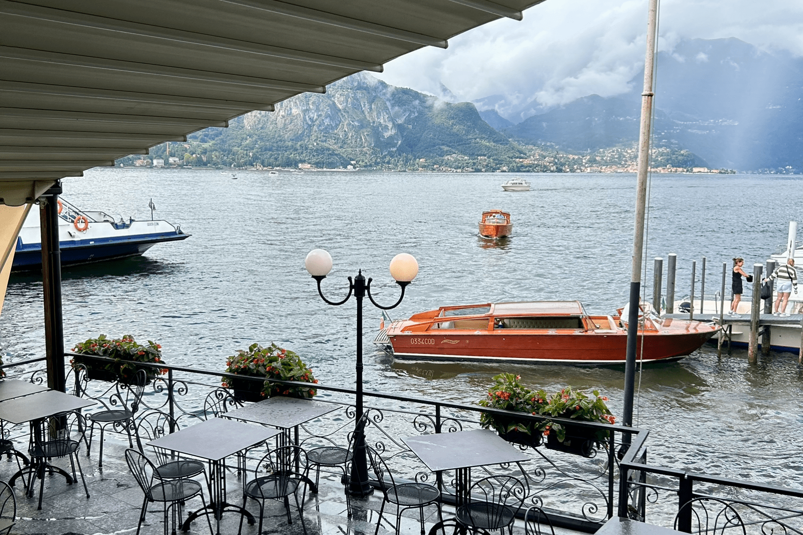 Lakeside café in Bellagio, Lake Como, Italy, with outdoor tables under a canopy, wooden boats docked at a pier, and misty Alps in the background on a rainy day.