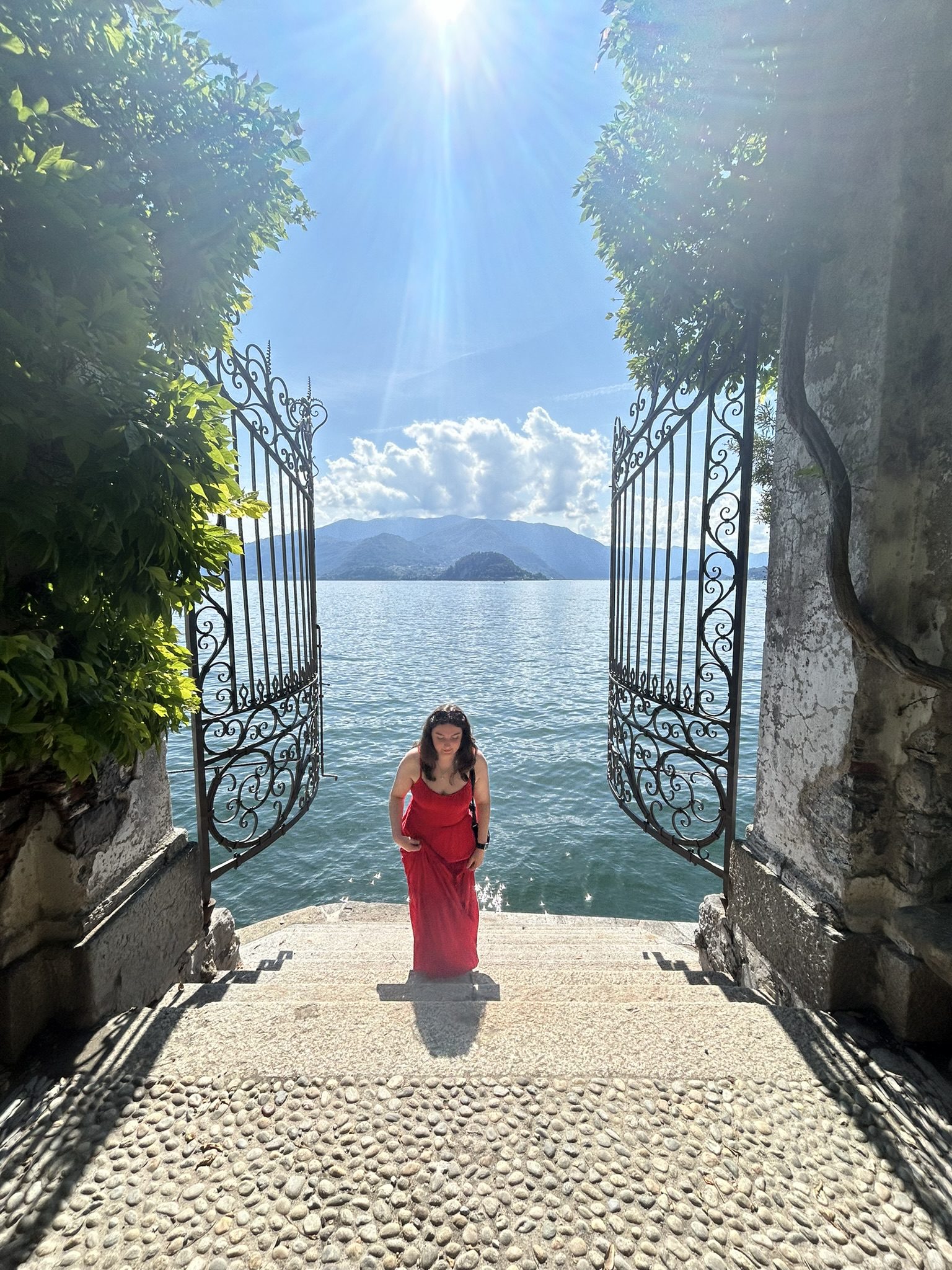 Woman in red dress on stone steps at Villa Cipressi, Lake Como, framed by iron gates with lake and mountain view.