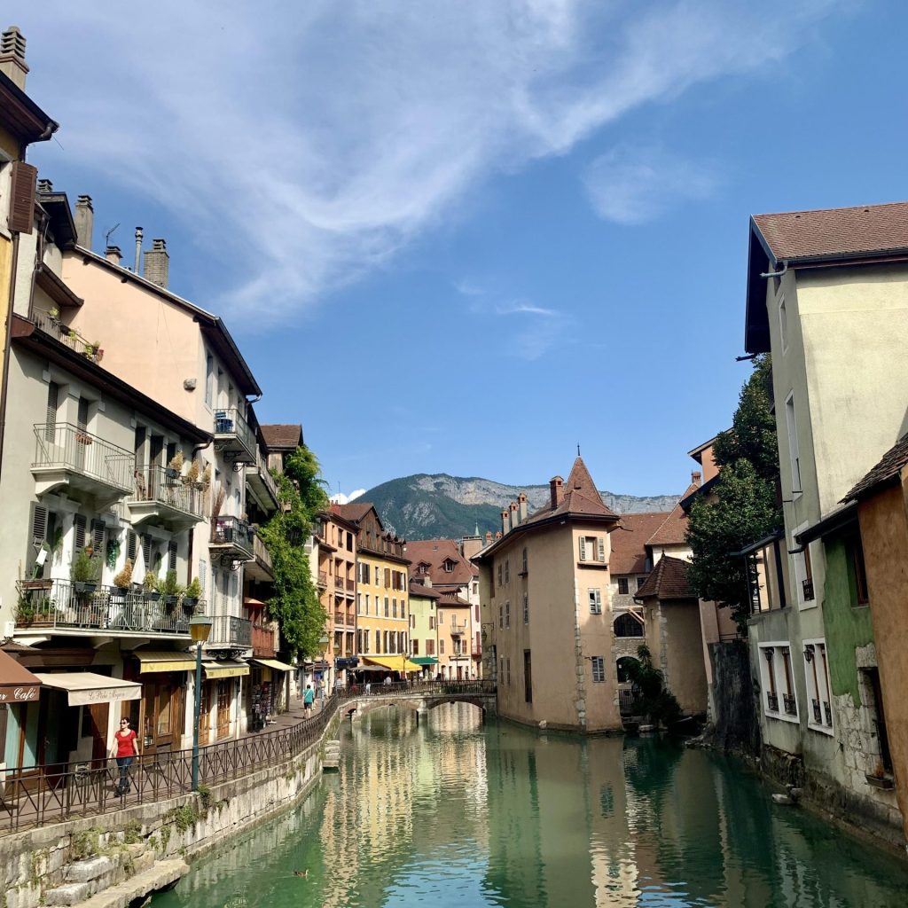 Scenic canal view in Annecy, France, with colorful historic buildings lining the waterway, a small stone bridge in the distance, and mountains in the background under a blue sky.