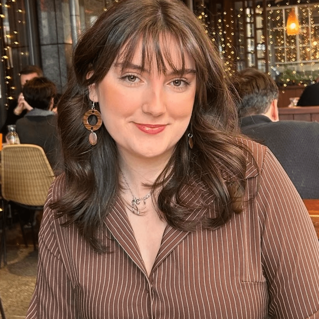 Girl in a brown striped shirt smiling in a cozy London restaurant, with wooden decor, string lights, and wicker lamps in a warm dining atmosphere.