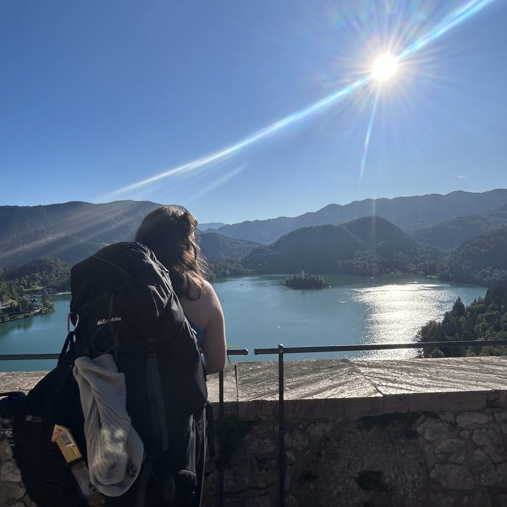Female hiker with backpack looking over a stone ledge at Lake Bled, Slovenia, with Bled Island and mountains under a sunny sky with lens flares.