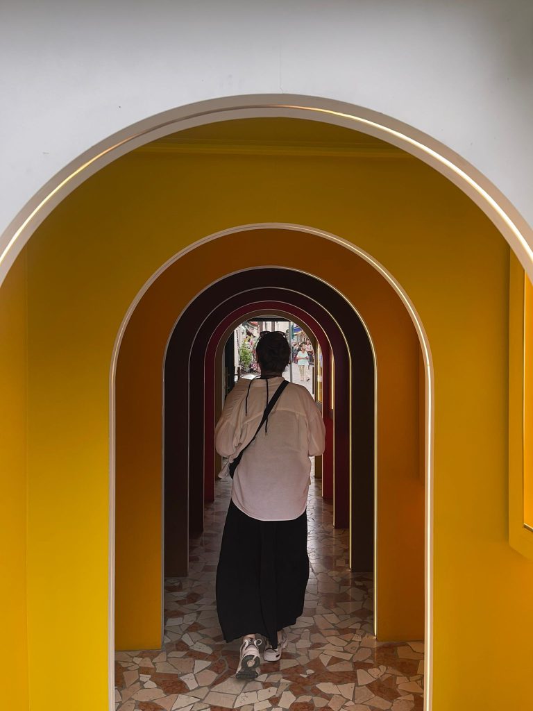 Girl walking through a multi coloured tunnel, with the primary colour being yellow. The tunnel is located in Paris. 