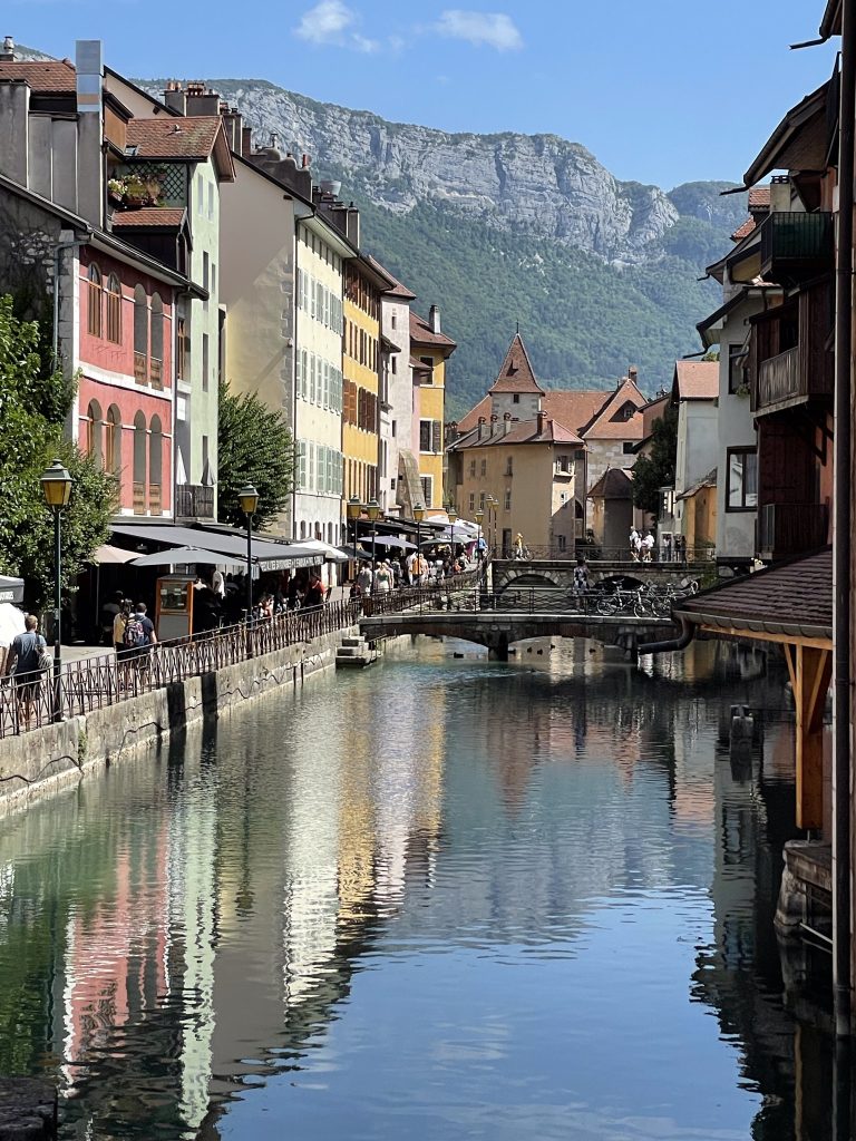 Colourful houses lining the side of the canal in Annecy with bridges over the canal and the French alps in the background. 