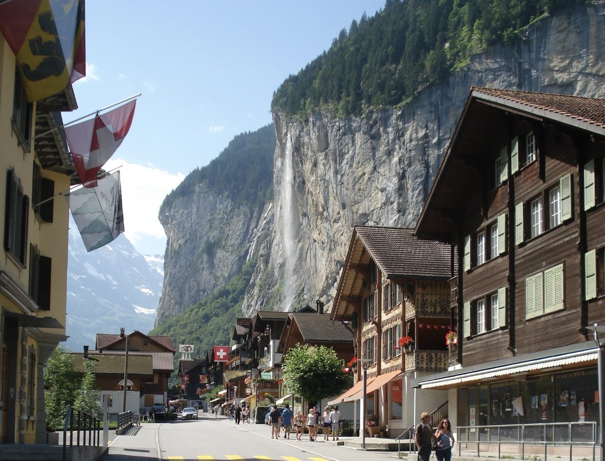 Charming street in Lauterbrunnen, Switzerland, with traditional Swiss chalets, Staubbach Falls cascading down a steep cliff, and snow-capped mountains under a clear blue sky.