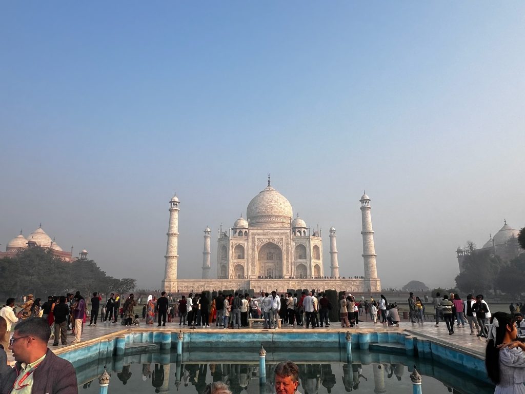 Taj Mahal in the daytime. Blue sky and crowds, with the reflecting pool in the foreground of the image. 